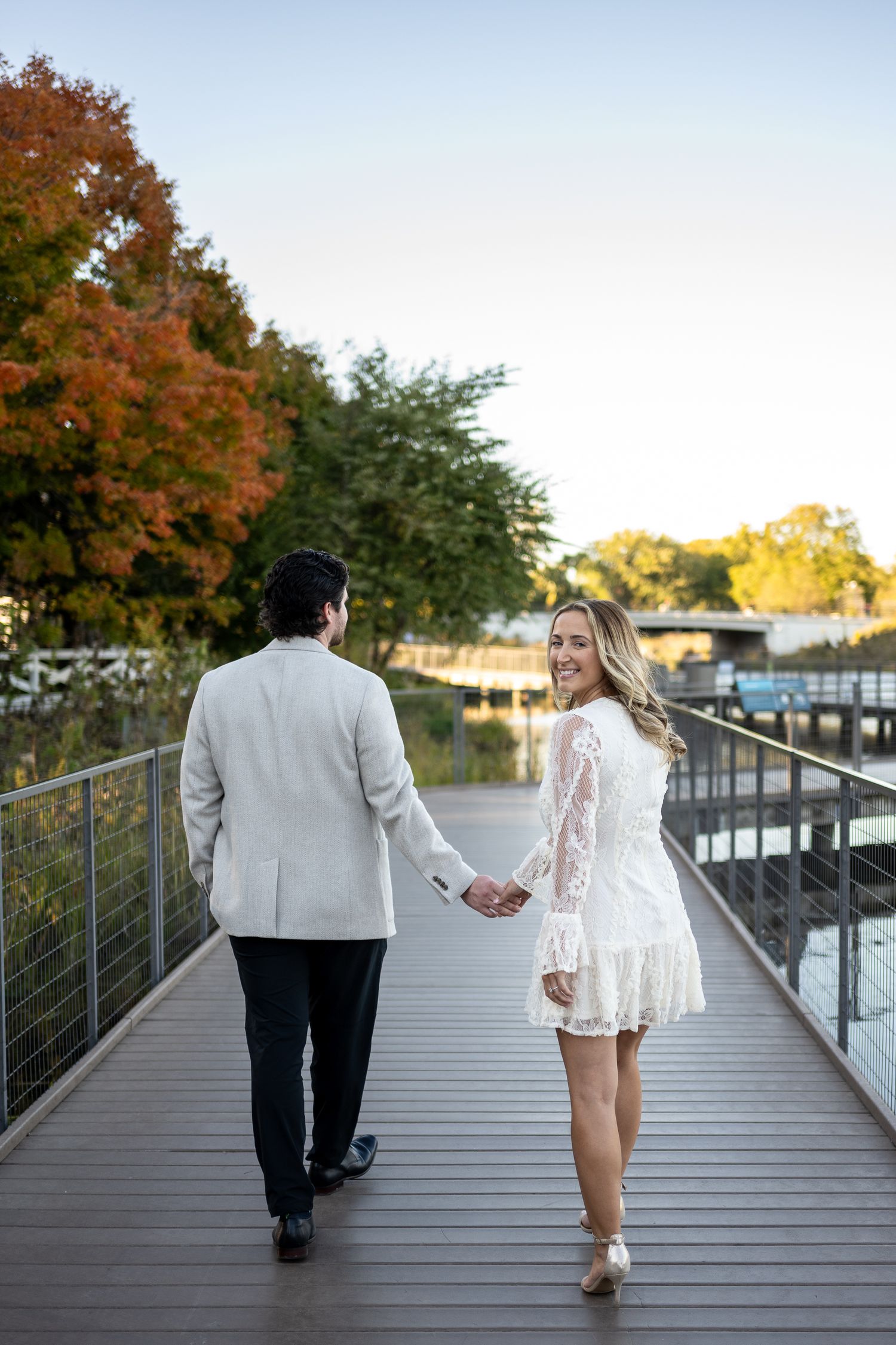 Couple holding hands walking on a boardwalk in autumn, woman in white lace dress smiling back at camera.