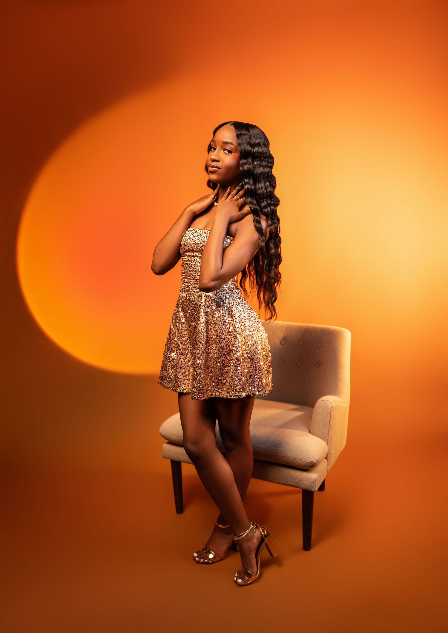 Young woman in gold sequin mini dress and heels poses elegantly beside a chair against warm orange backdrop.