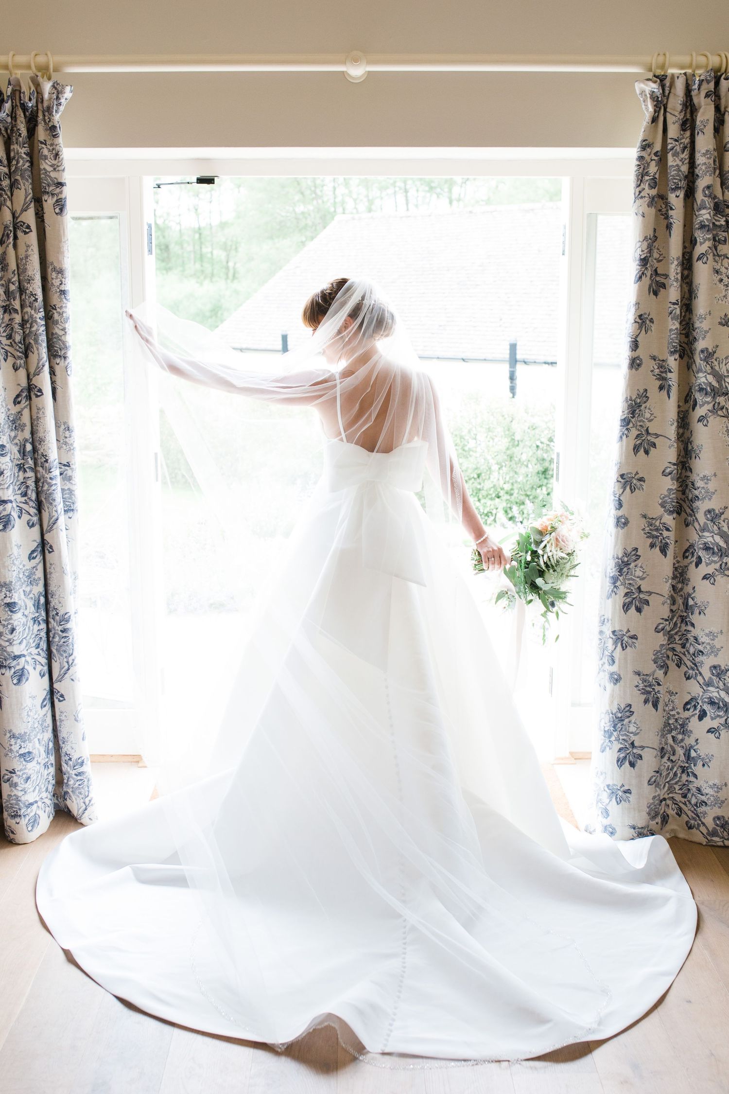 Bride in white gown and veil stands before sunlit window holding bouquet, cathedral train spread elegantly on floor Wilsons Coastal Resort