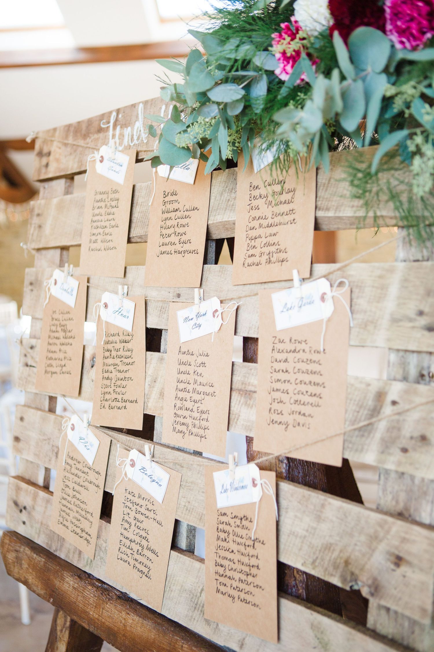 Rustic wooden pallet wedding seating chart with kraft paper cards, clothes pins, and floral arrangement on top.