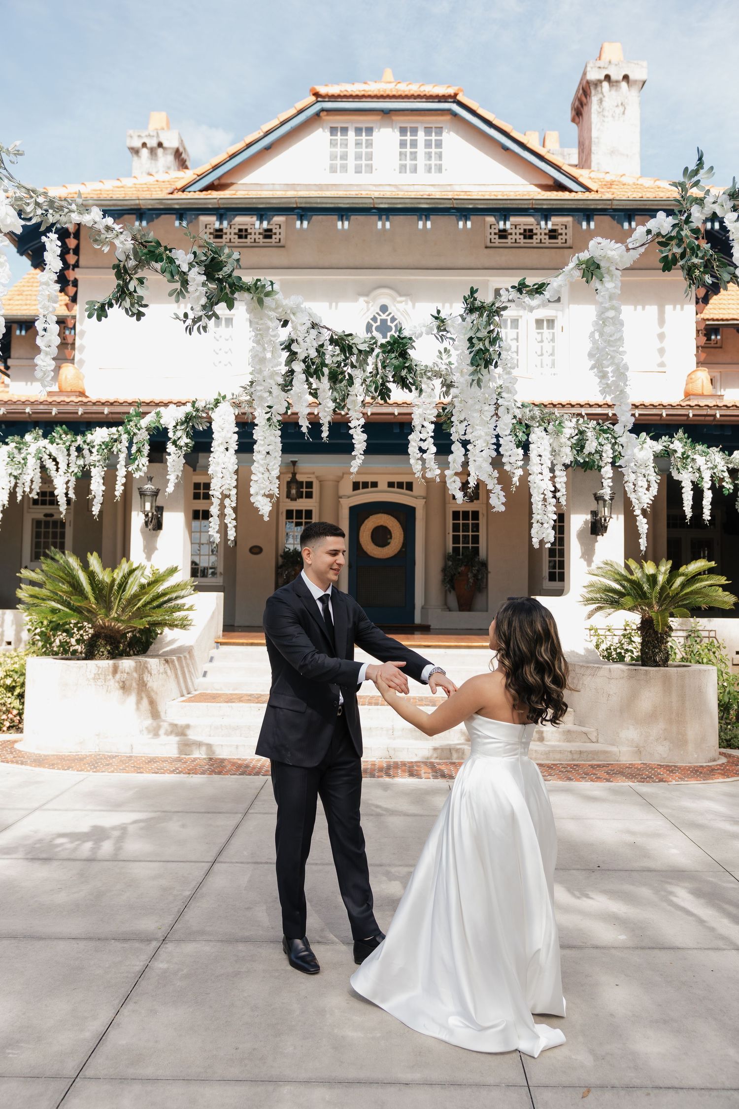 Bride and groom dancing outside an elegant mansion adorned with hanging white wisteria flowers on a sunny day.