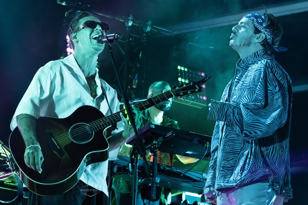 Two male musicians performing live on stage, one playing acoustic guitar and singing, both smiling under concert lights.