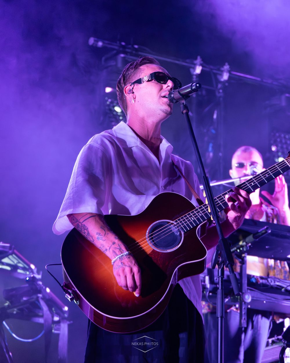 Tattooed musician playing acoustic guitar on stage under purple lights at live concert.