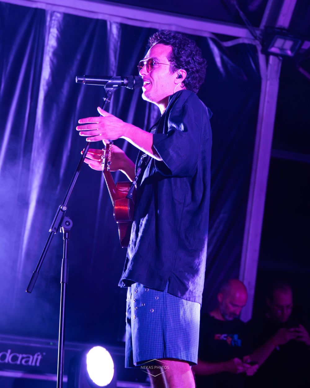 Male singer performing on stage with microphone and guitar under purple lights at outdoor music festival.