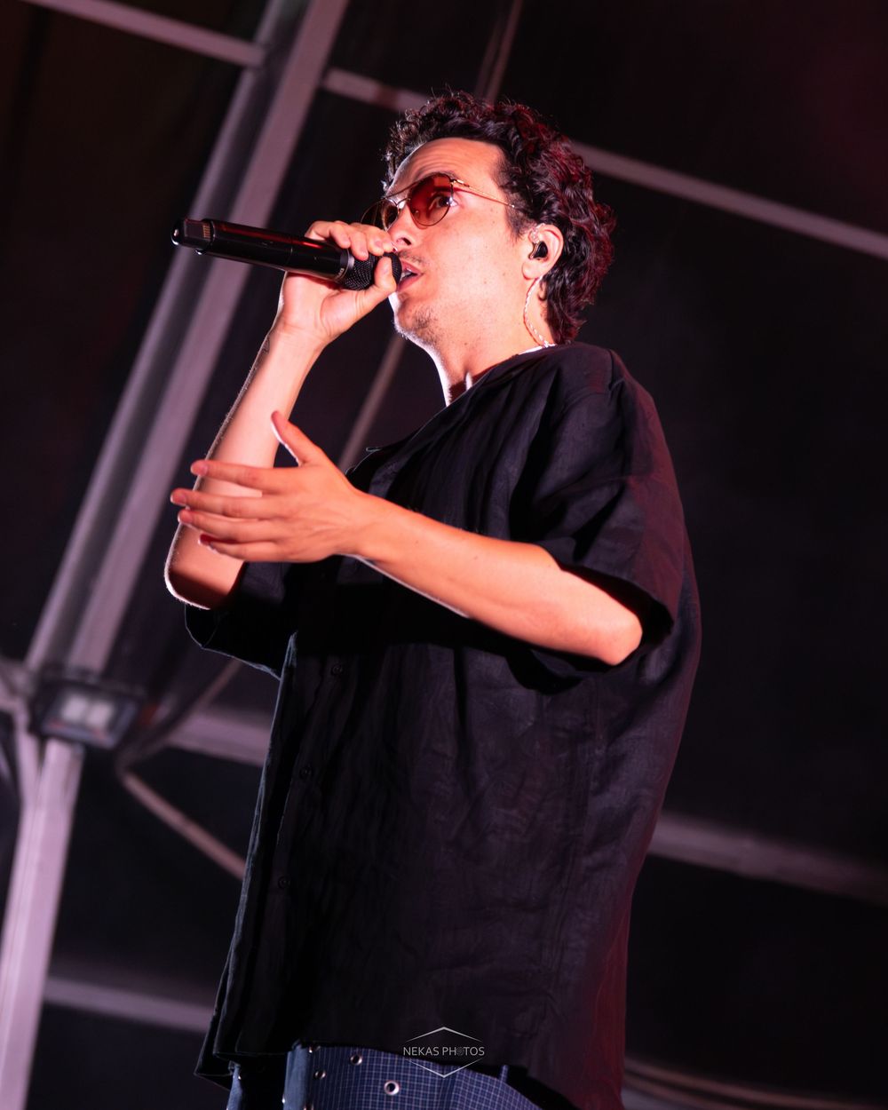 A male singer performs on stage wearing black oversized shirt and sunglasses, holding a microphone close to his mouth.