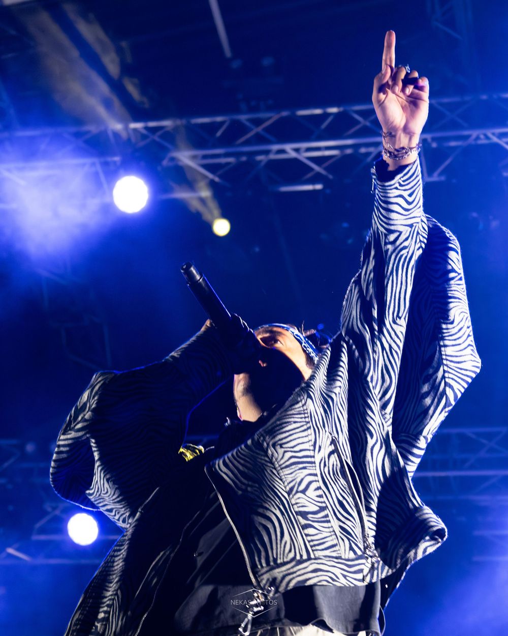 A performer in a zebra-print jacket raises one finger skyward while holding a microphone on a blue-lit concert stage.