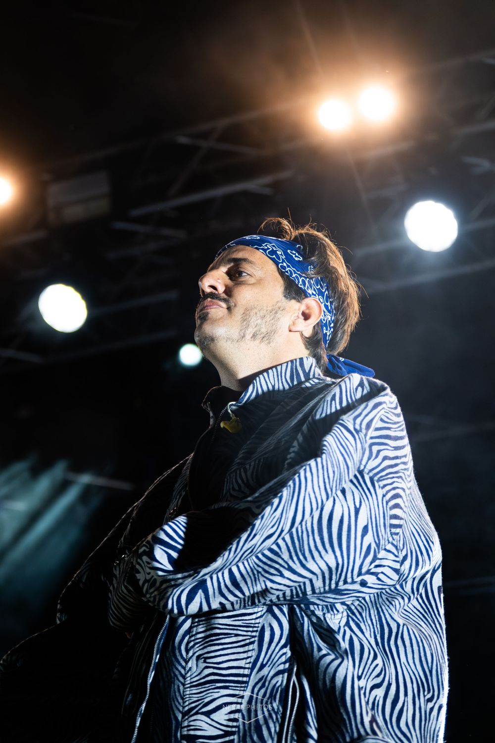 Man in zebra-print jacket and blue bandana performing on stage under bright concert lights.