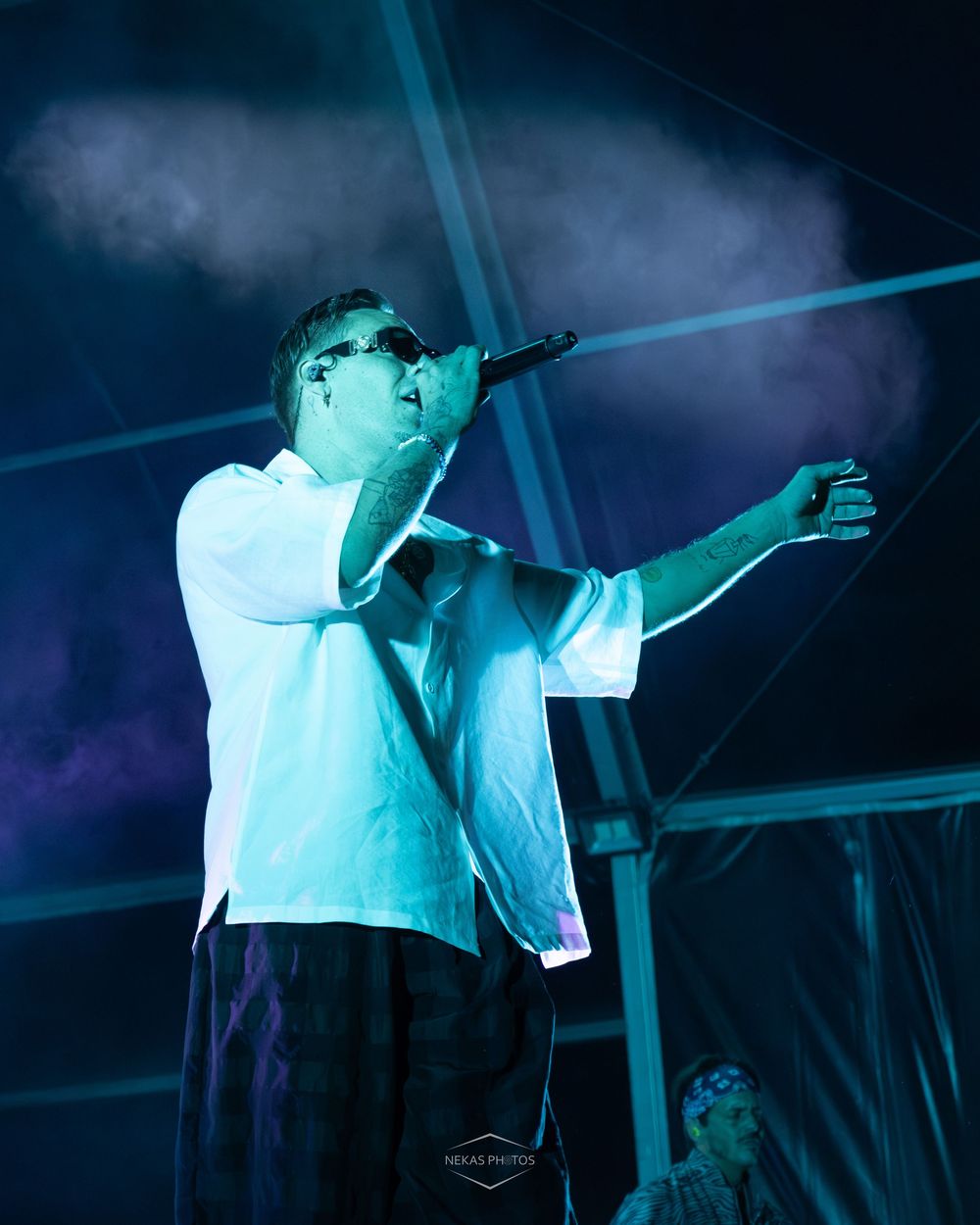 A male performer in a white shirt and sunglasses sings into a mic on a blue-lit outdoor stage.