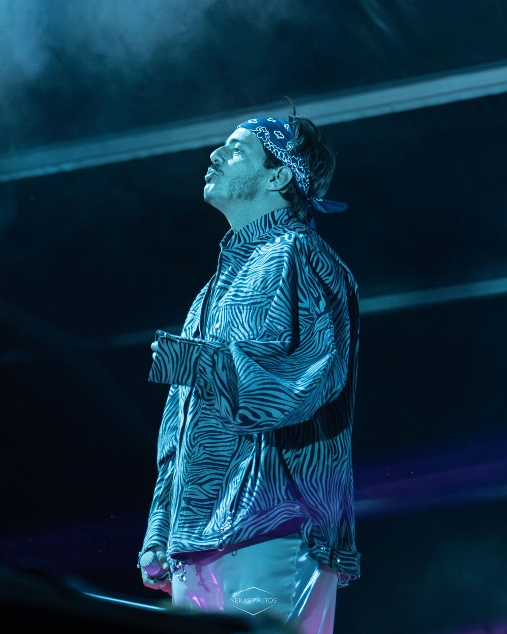 Male performer in zebra-print jacket and bandana on stage under blue concert lighting, looking upward dramatically.