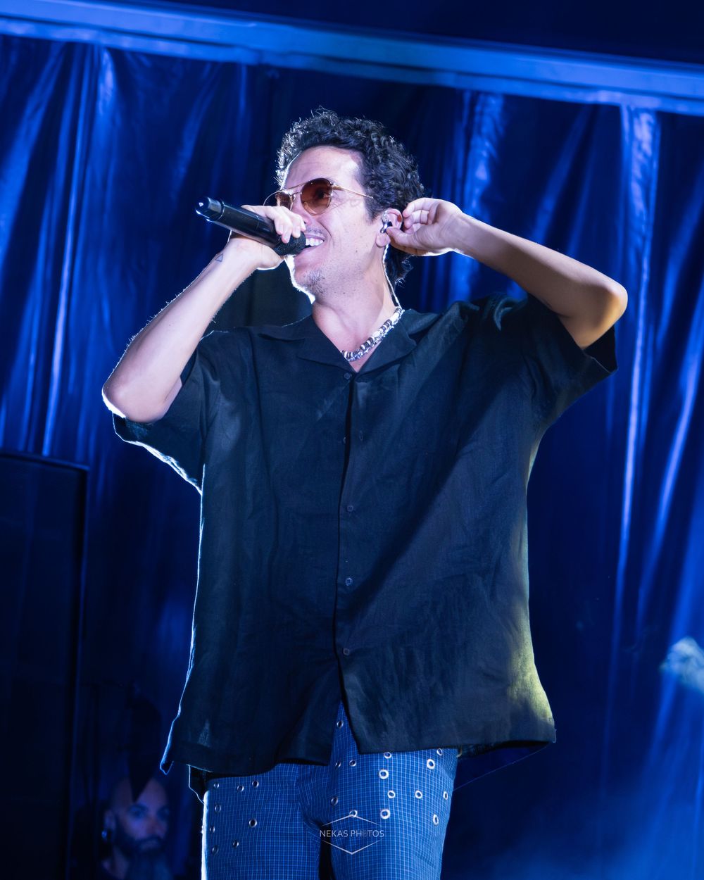 Male singer performing on stage in black shirt and sunglasses, holding microphone under blue stage lighting.