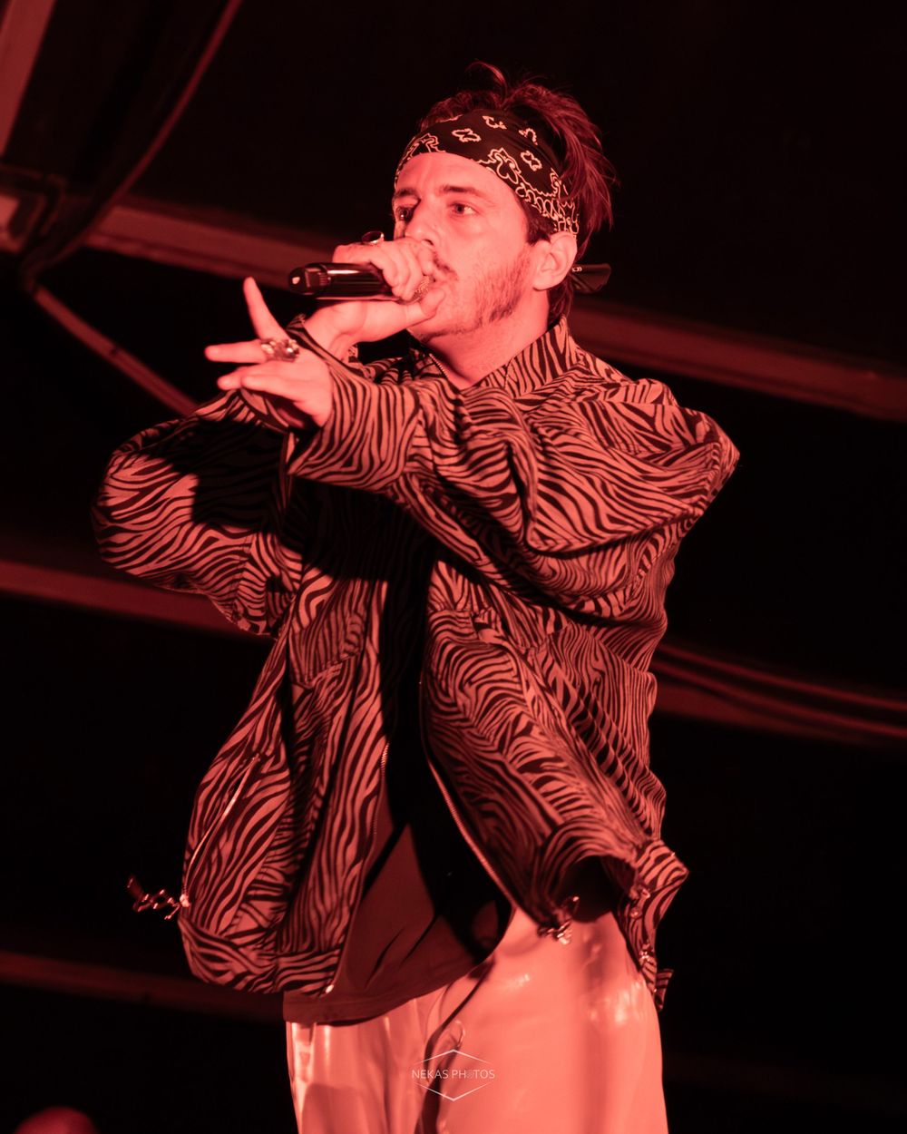 Male rapper performing on stage with microphone, wearing zebra print jacket and bandana headband under red stage lighting.