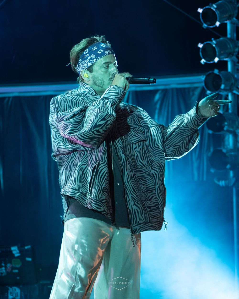 Male rapper performing on stage wearing zebra print jacket and blue bandana headband, holding microphone under blue stage lights.
