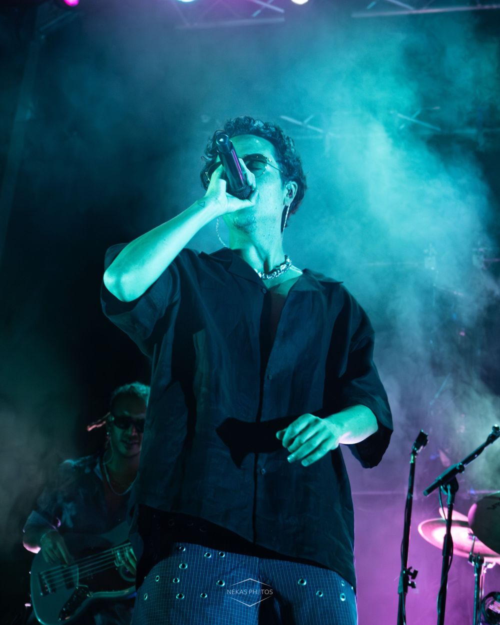 Male singer performing on stage under teal lighting with microphone, wearing black shirt, smoke effects in background.