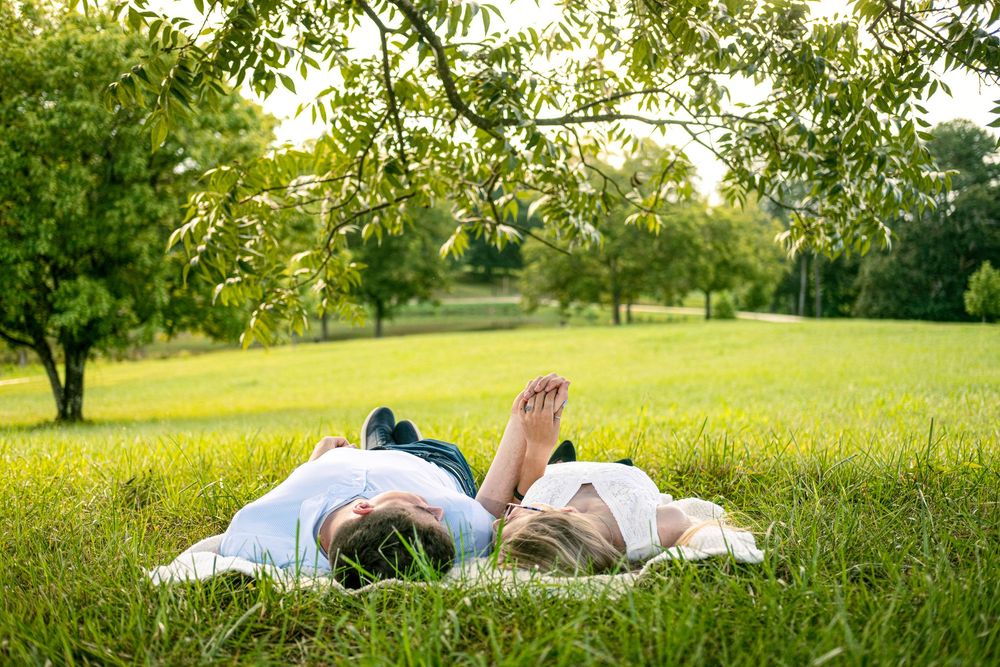 Two people lying on lush green grass under a tree in a sunny park, relaxing together on a warm summer day.