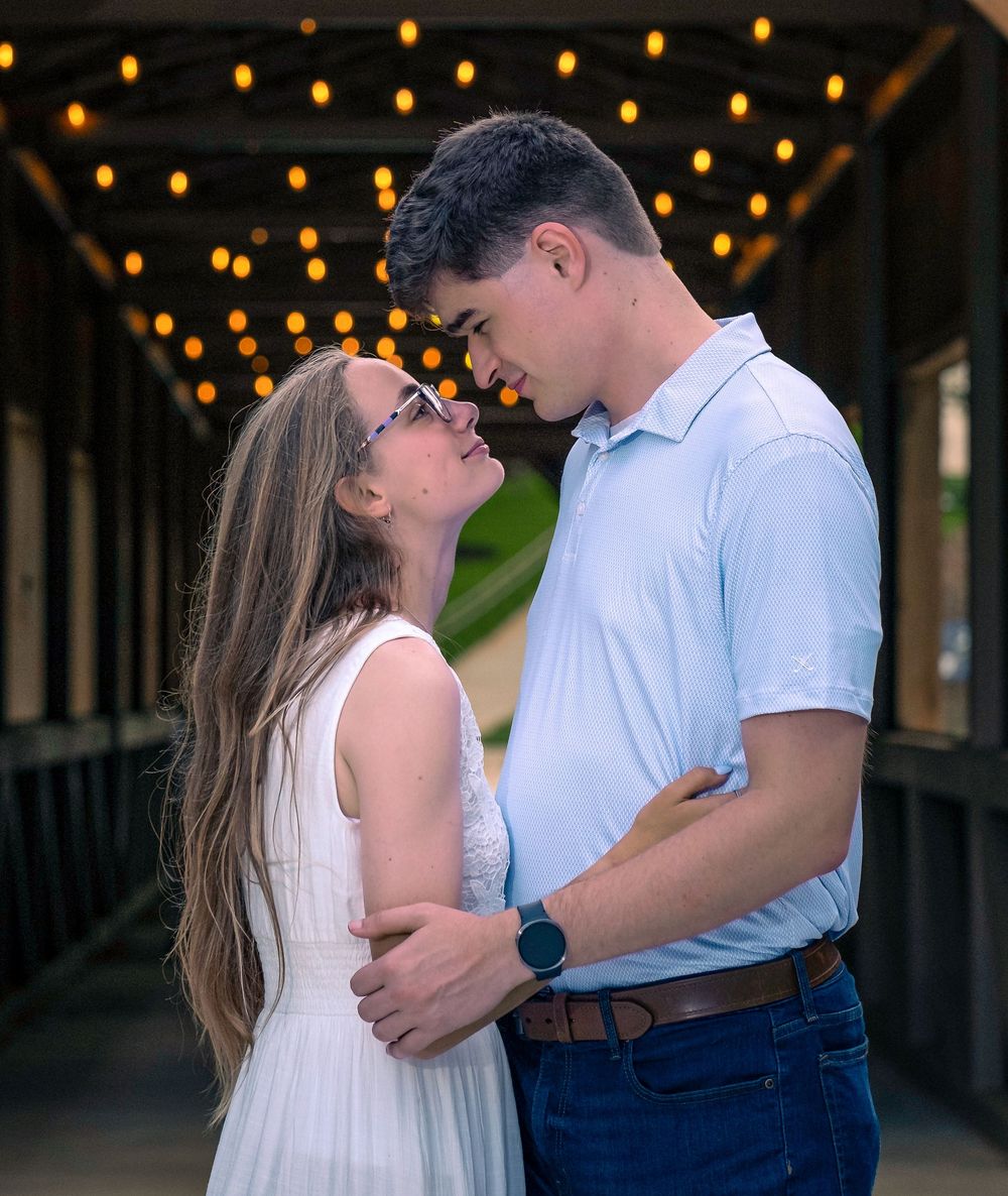 Couple sharing a romantic moment on a bridge with string lights, woman in white dress and man in blue shirt and jeans.