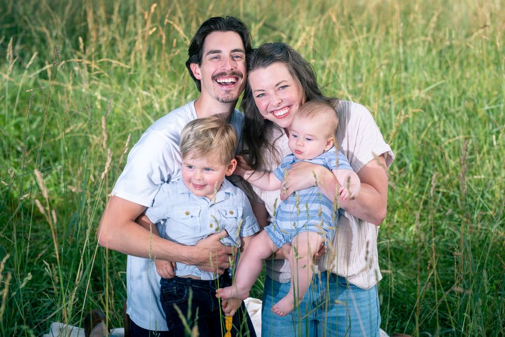 Happy young family with two toddlers smiling outdoors in a lush green field during a summer photo session.