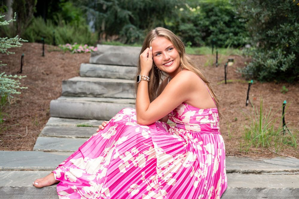 Young woman in pink floral dress sitting on stone steps in a garden, smiling for senior portrait photo.