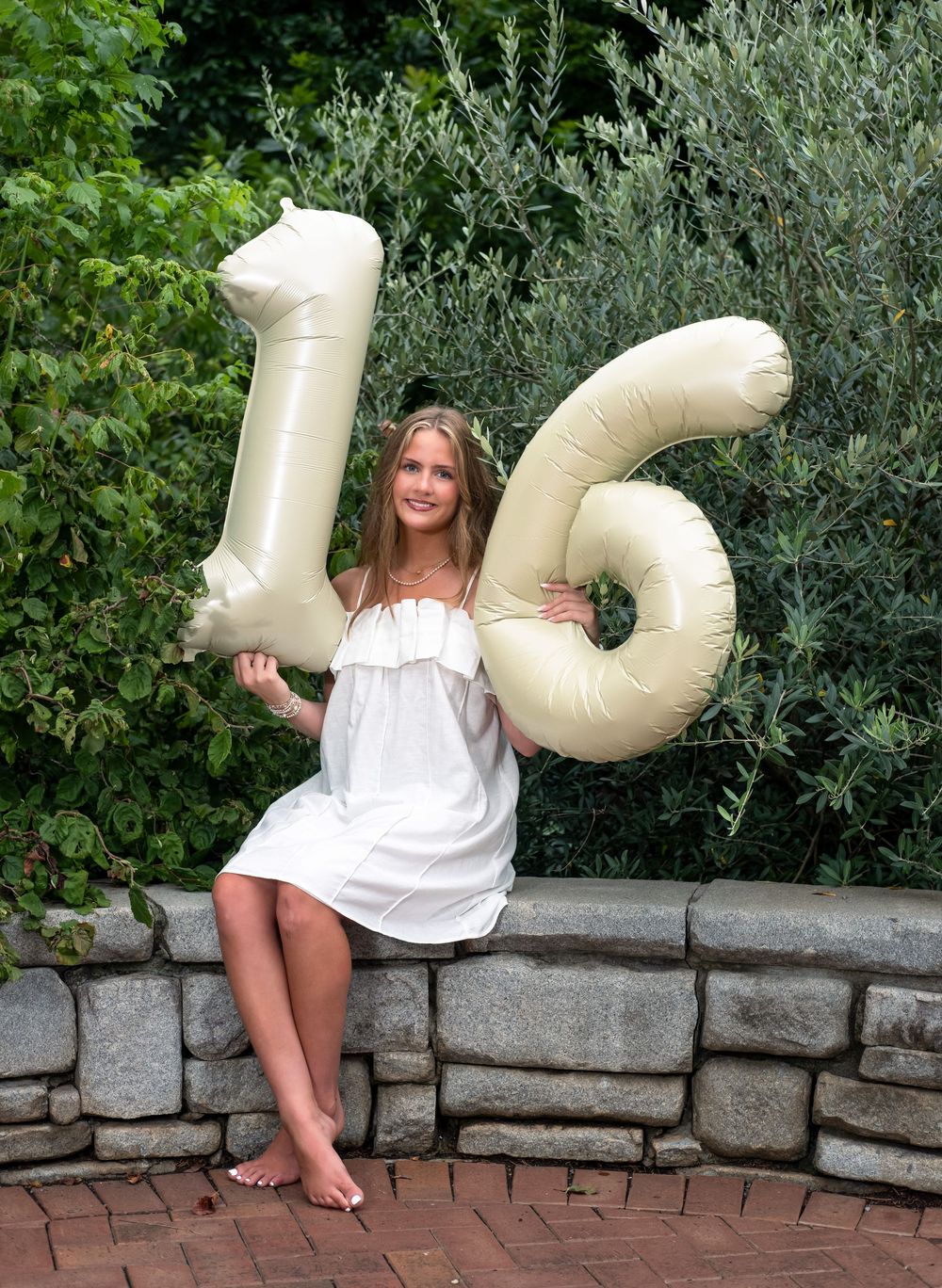 A smiling girl in a white dress holds large cream-colored number 16 balloons while sitting on a stone wall outdoors.