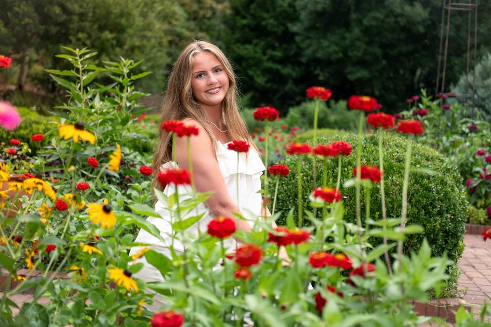 Young blonde woman in white dress smiling in a vibrant garden with red zinnias and lush green plants.