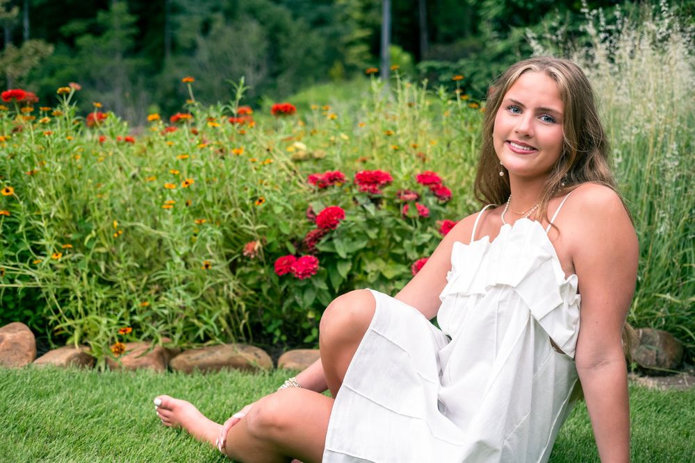 Young woman in white dress sitting on green grass in front of vibrant red and orange flower garden.