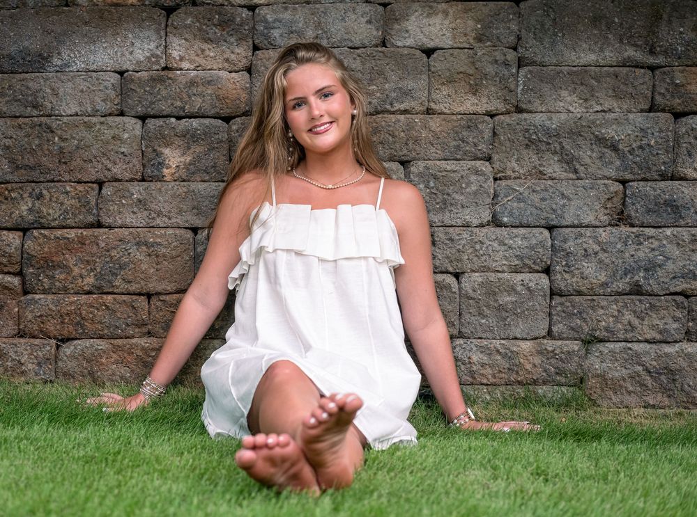 Young woman in white sundress sitting barefoot on green grass against rustic stone wall outdoors.
