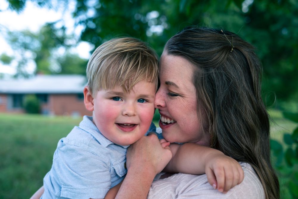 A smiling toddler boy hugs a laughing brunette woman outdoors with green trees in the background.