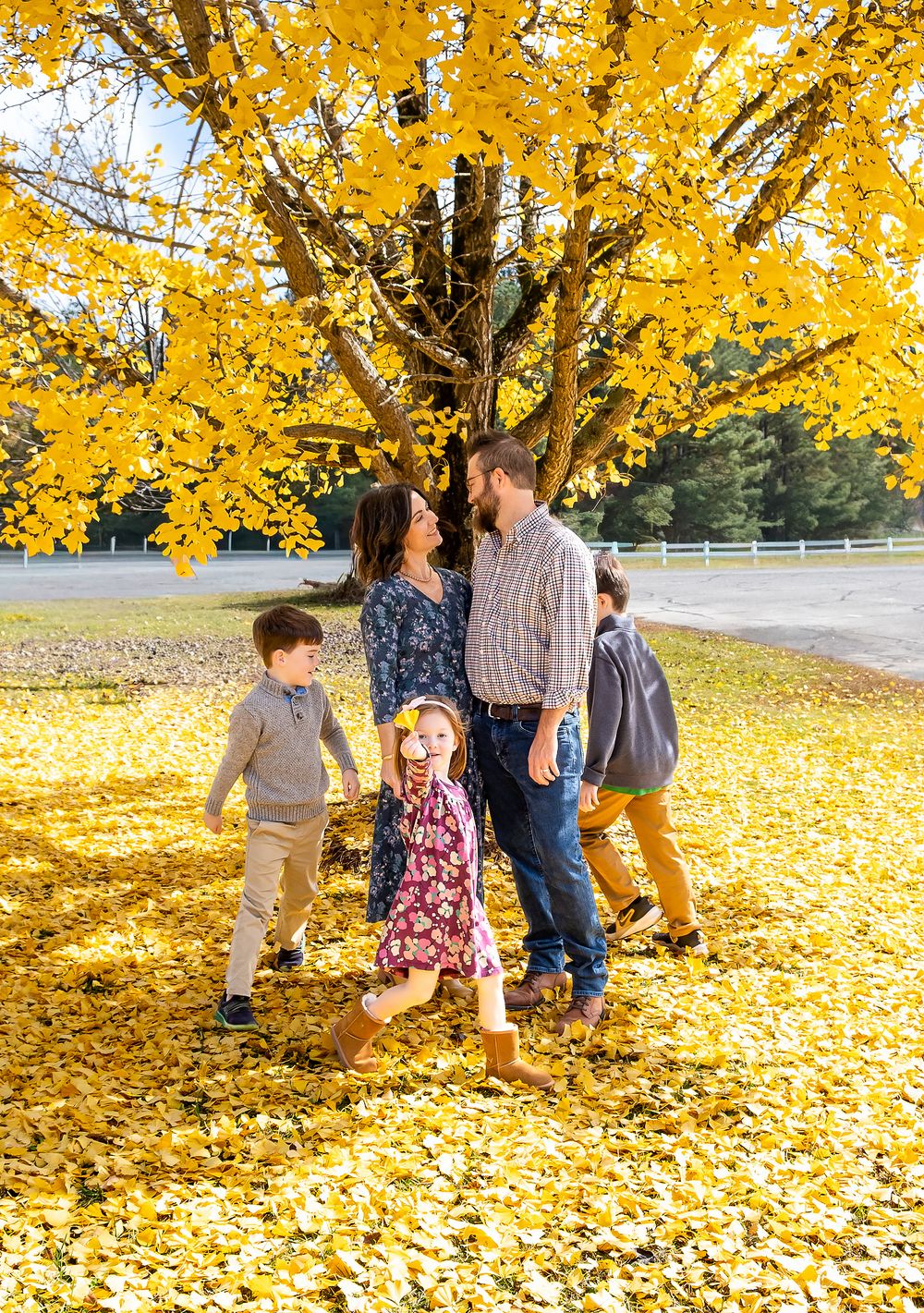 Family of five playing in golden fallen leaves under a vibrant yellow autumn tree outdoors.