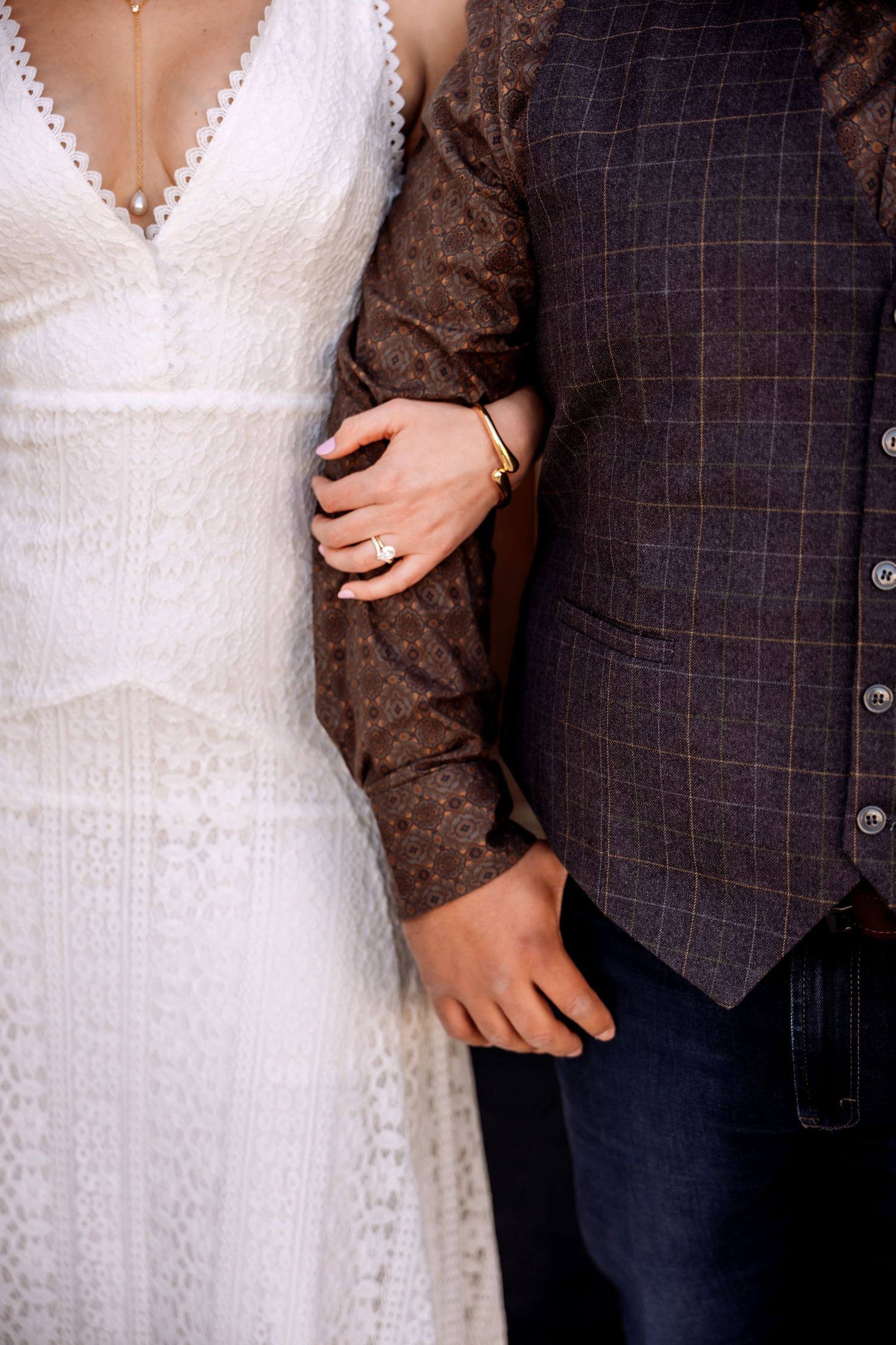 A close-up wedding portrait of a bride in a lace gown linking arms with a groom in a textured vest, highlighting the refined details of thei