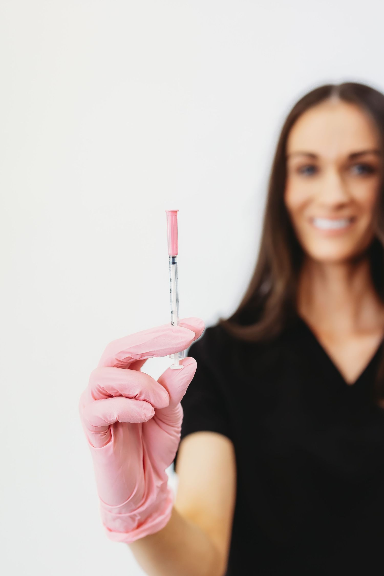 Smiling female aesthetician in pink gloves holding a medical syringe for cosmetic injection treatment.