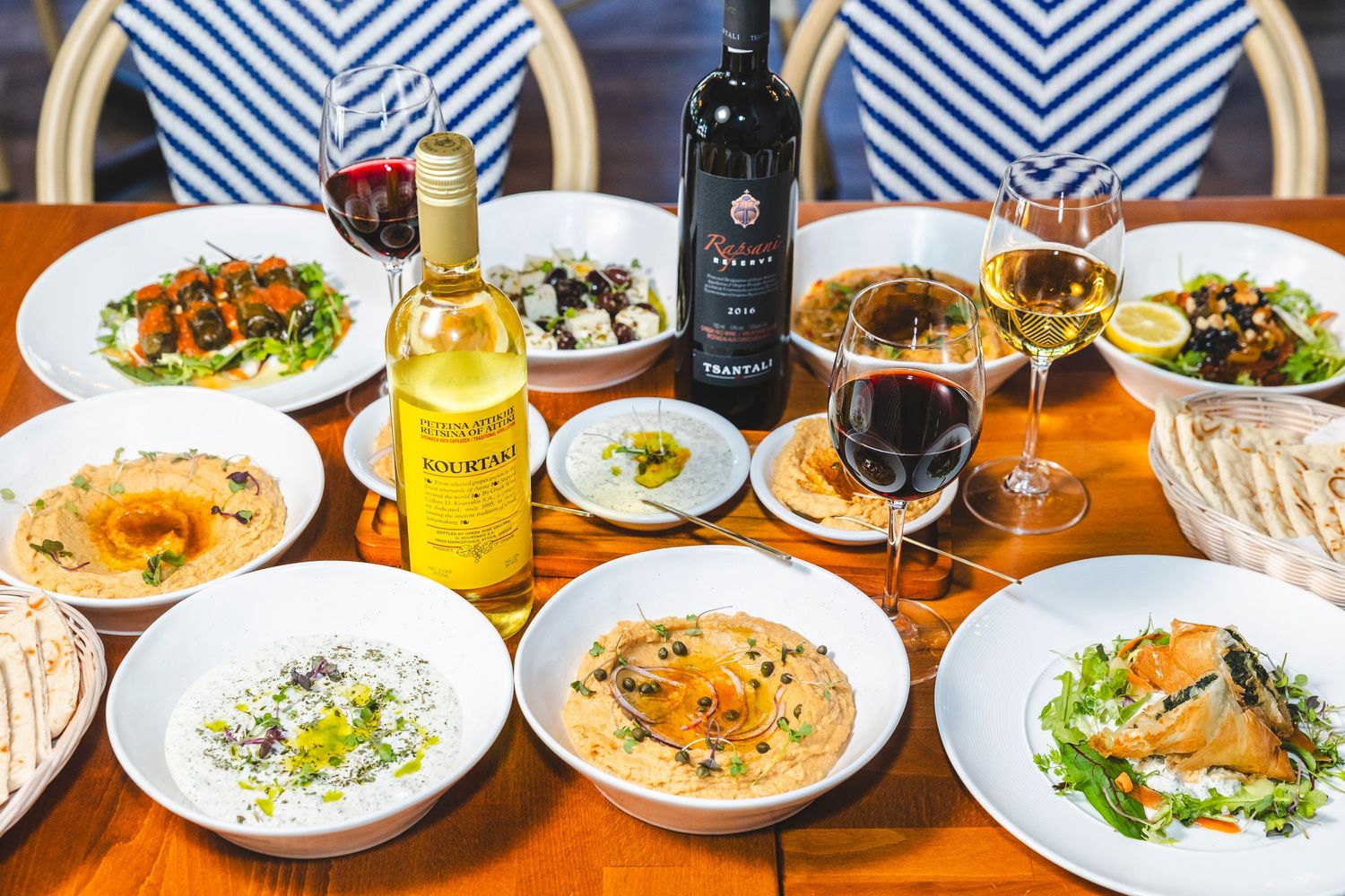 A spread of Mediterranean dishes with hummus, salads, and wine bottles on a wooden restaurant table.