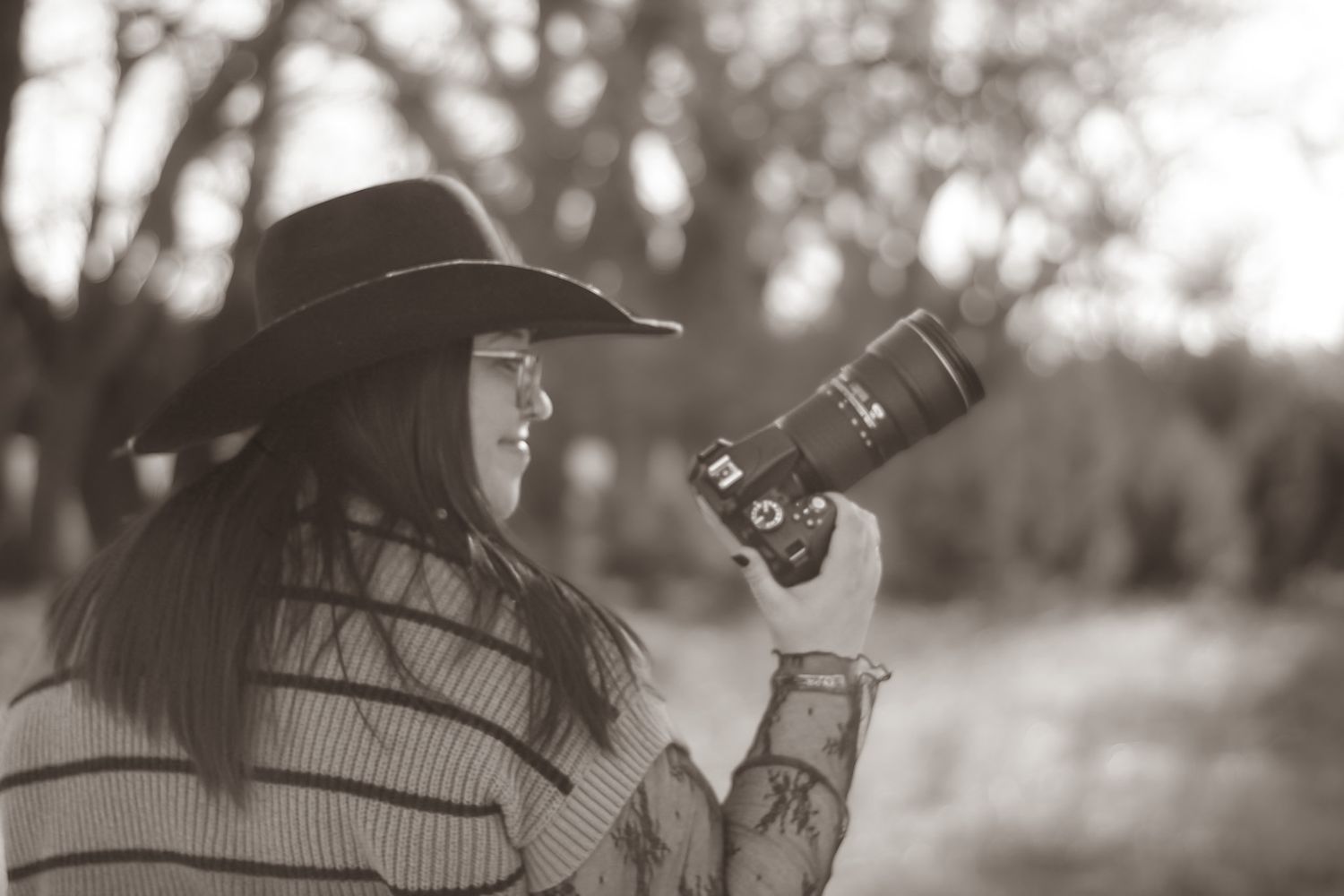 Black and white photo of a woman in a cowboy hat holding a DSLR camera outdoors among trees.