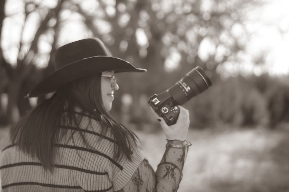 Black and white photo of a woman in a cowboy hat holding a DSLR camera outdoors among trees.