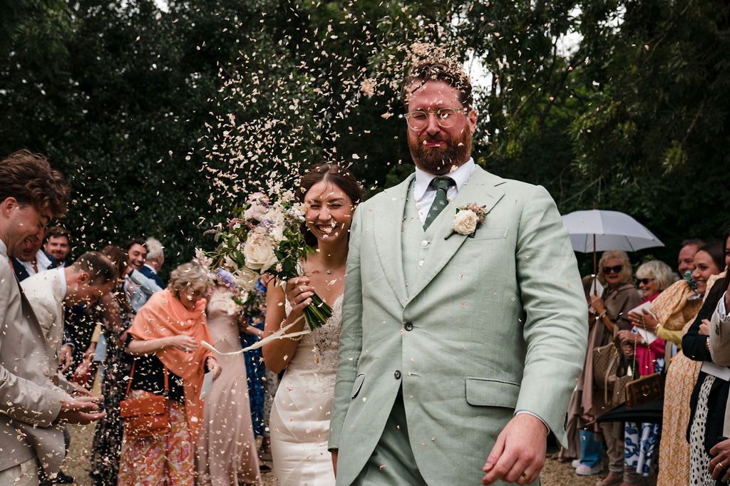 Bride and groom walk through confetti shower, groom in mint suit, bride in white dress, guests celebrating outdoors.