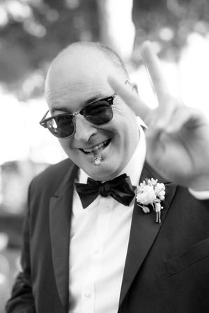 Groom in tuxedo and sunglasses flashing peace sign with boutonniere at wedding celebration.