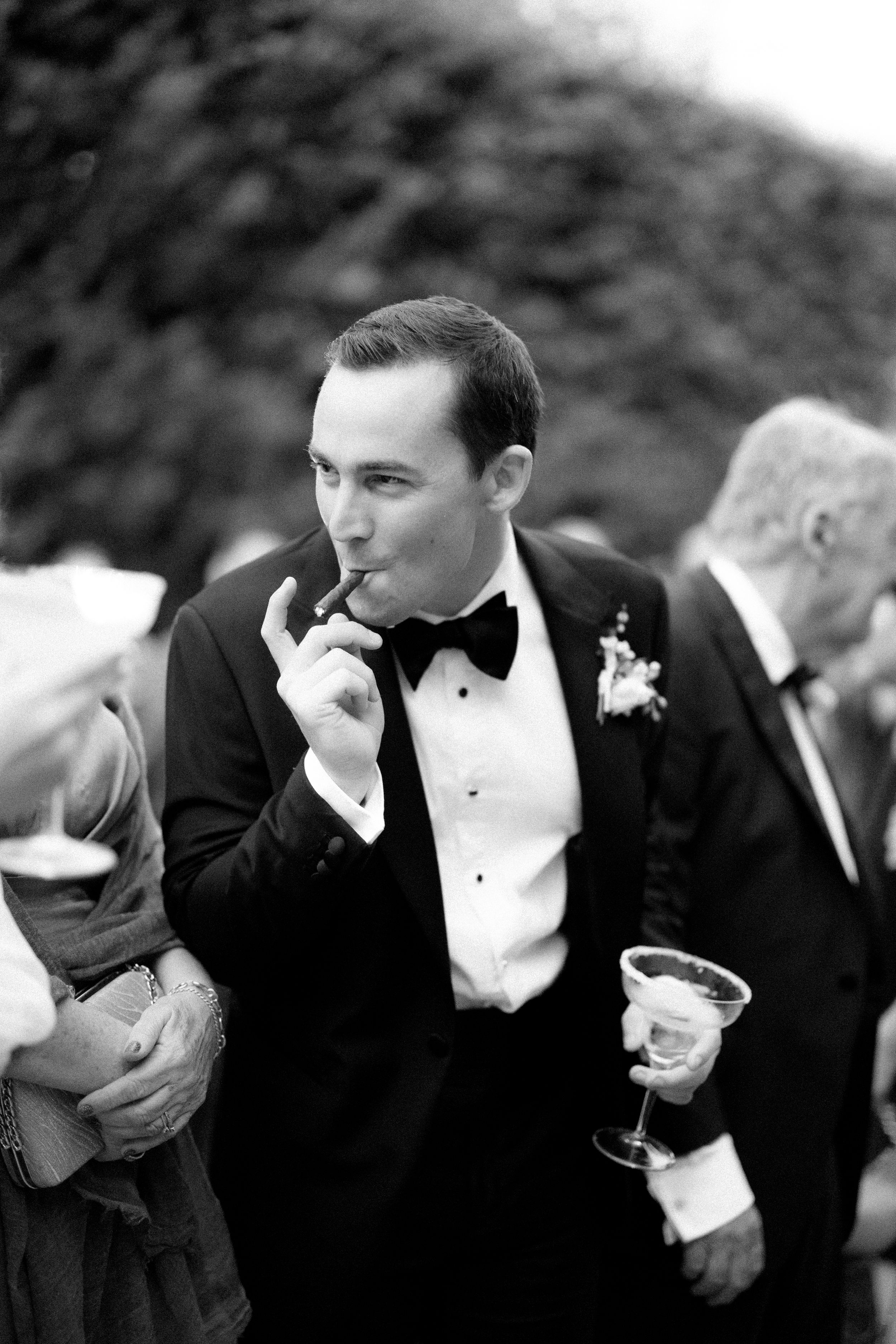 Groom in tuxedo smoking cigar and holding martini glass at elegant black and white wedding reception.