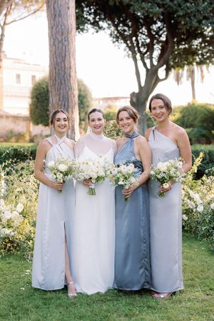Bride poses with three bridesmaids in mismatched grey gowns holding white bouquets in a garden.
