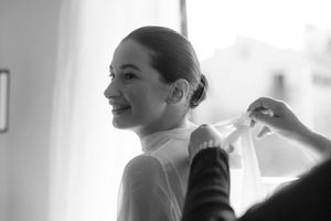 Black and white photo of a smiling bride having her wedding veil attached by a helper.