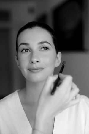 Black and white portrait of a bride having makeup applied with a brush, smiling softly in a white robe.