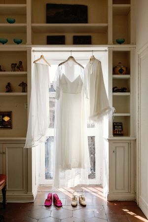 White wedding dress and bridal robes hanging in a sunlit built-in wardrobe with pink and nude bridal shoes below.