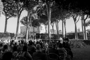 Black and white outdoor gathering with crowd seated among tall pine trees near a historic Italian villa.