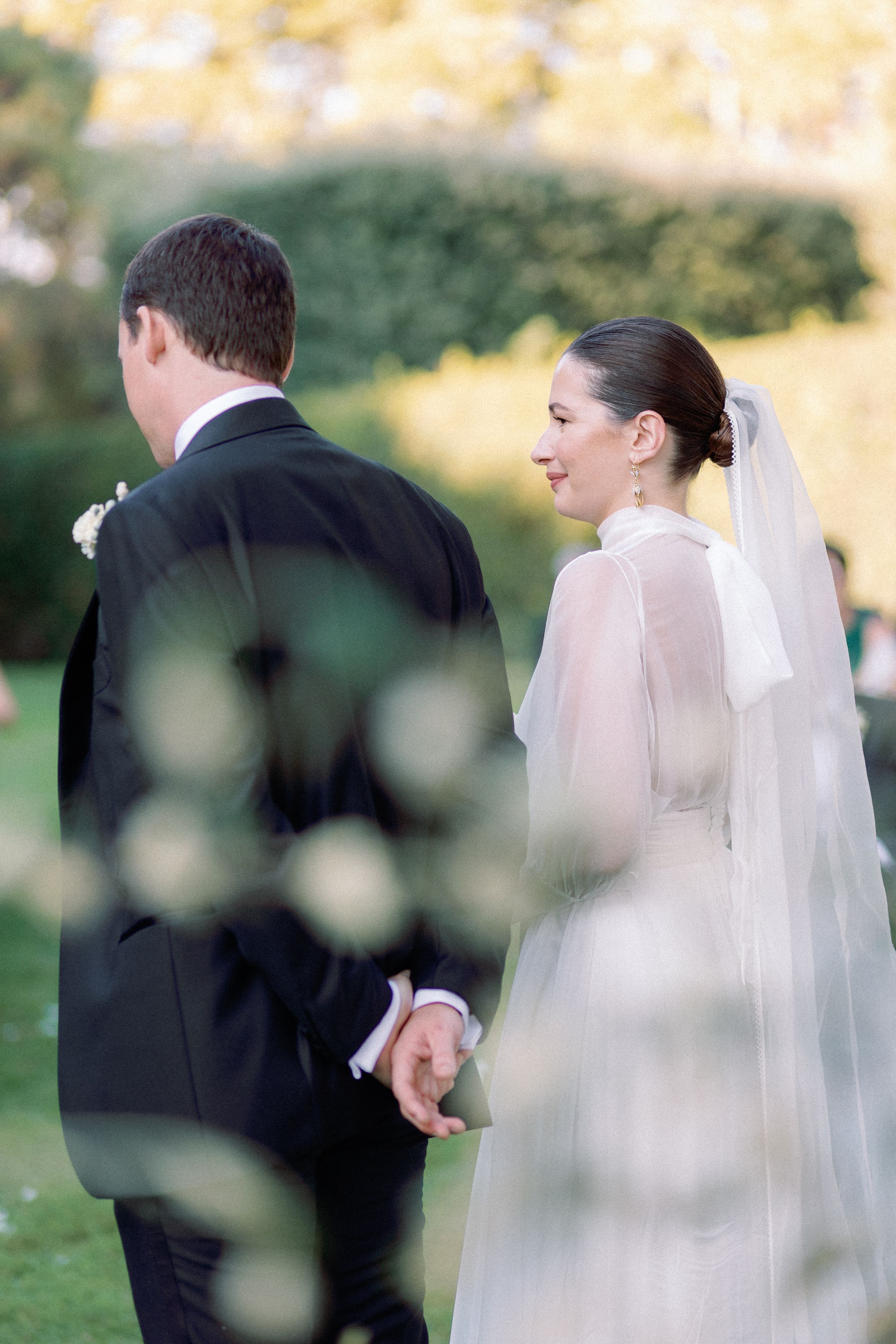 Bride in sheer white gown and veil holds groom's hand outdoors, smiling softly in a lush green garden setting.
