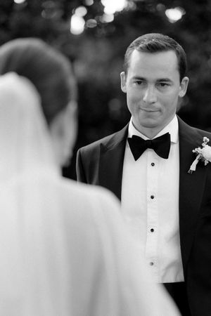 Groom in black tuxedo gazing at bride during outdoor wedding ceremony, black and white photo.