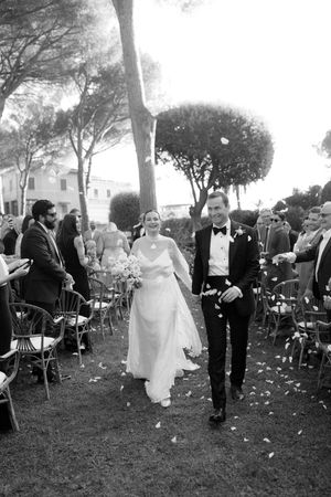 Bride and groom walk down outdoor aisle in black and white wedding photo with guests and trees.