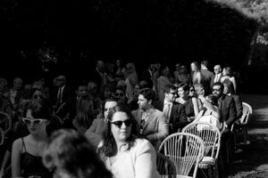 Black and white photo of guests seated outdoors at a garden wedding ceremony, wearing sunglasses in bright sunlight.