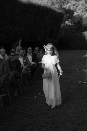 Flower girl in white dress and floral crown walks down outdoor wedding aisle carrying a wicker basket.