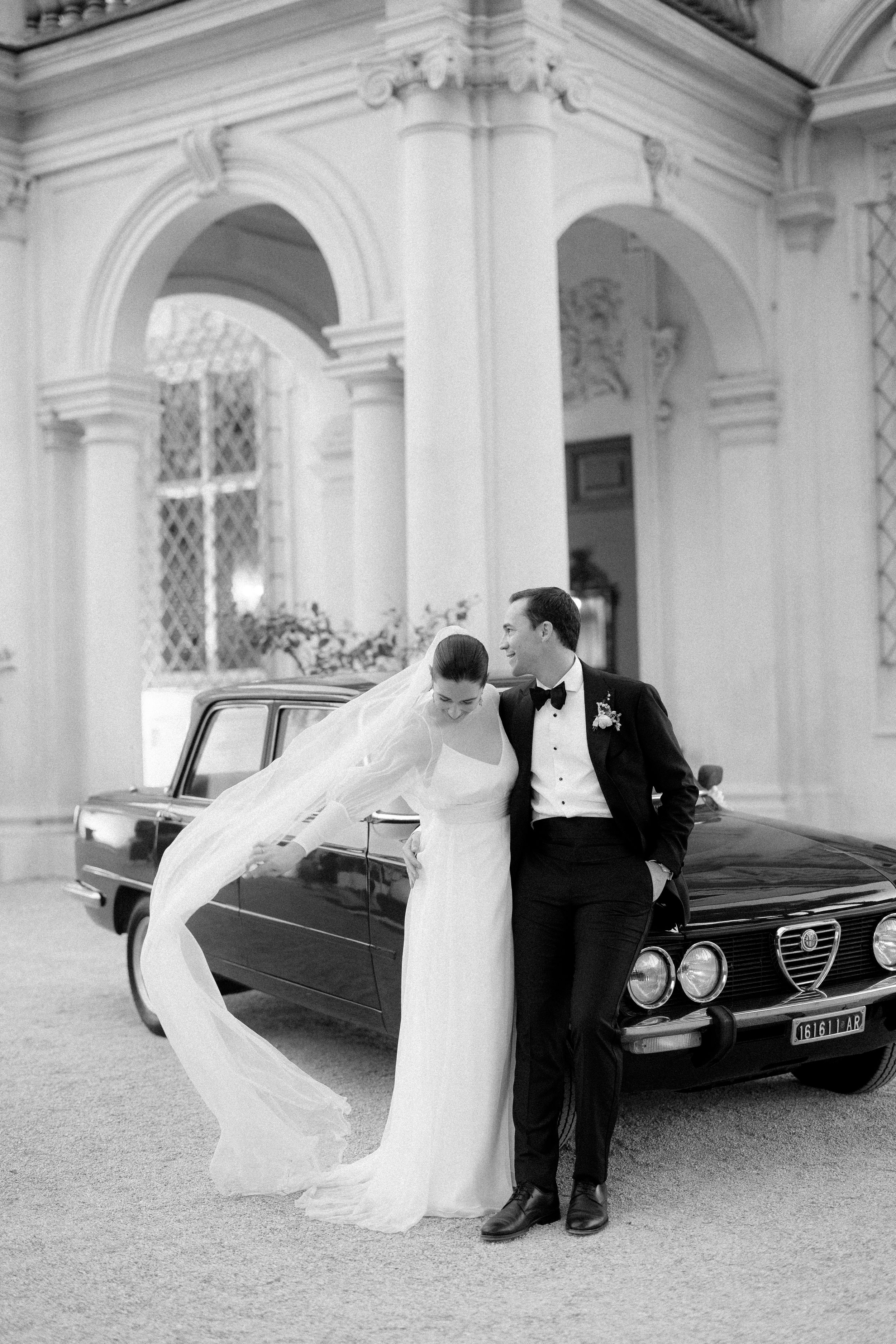 Bride and groom pose beside vintage Alfa Romeo car in front of ornate classical architecture.