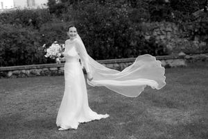 Black and white photo of a bride in a flowing white gown and veil billowing in the wind, holding a bouquet outdoors.