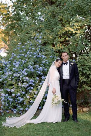 Bride in white gown with long veil leans on groom in black tuxedo before blue flowering bushes.