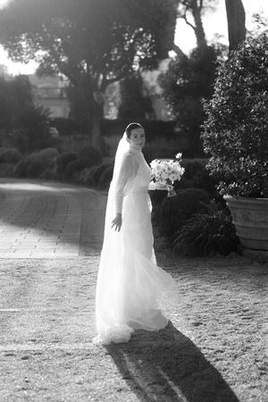 Elegant bride in flowing white gown and veil holds bouquet while posing in sunlit garden setting.