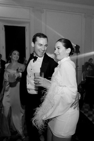 Couple dancing at formal event, man in tuxedo holding drink, woman in white feathered outfit, black and white photo.